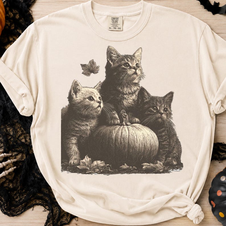 May include: A cream-colored short-sleeve t-shirt featuring a dark brown graphic print of three kittens sitting around a large pumpkin. The kittens are surrounded by scattered autumn leaves. The shirt has a crew neckline and a Comfort Colors brand label.