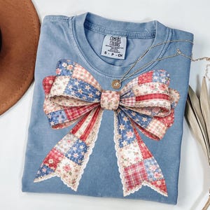 Könnte beinhalten: Ein blaues Comfort Colors T-Shirt mit einem großen Schleifendesign im Patchwork-Muster in Rot, Weiß und Blau. Die Schleife hat Blumen- und Sternmuster mit Spitzenbesatz. Eine goldene Halskette mit Anhänger liegt über dem Shirt.