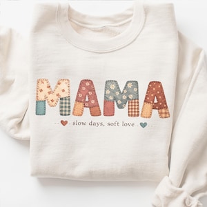 May include: Cream-colored sweatshirt with the word "MAMA" in patchwork letters. The letters are made of various patterned fabrics in shades of beige, pink, blue, and brown. Below the word are the words "slow days, soft love" with heart accents.