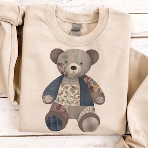 May include: A beige sweatshirt featuring a patchwork teddy bear design. The bear has a variety of fabric patterns, including floral, plaid, and solid colors in shades of blue, brown, and beige. The sweatshirt has a crew neck and long sleeves.