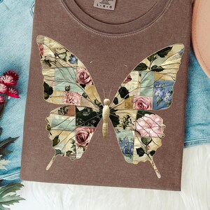 以下が含まれることがあります： 茶色のTシャツ。大きな蝶のデザインが特徴です。 蝶の羽は、ピンク、青、クリーム色のバラやその他の花を使った花のパッチワークで構成されています。 シャツはニュートラルカラーです。