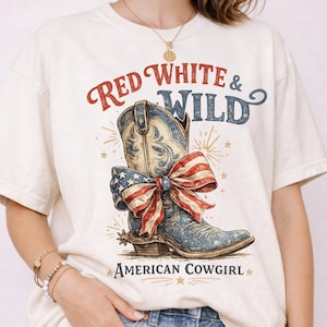 Puede incluir: Camiseta color crema con un gráfico de una bota de vaquero azul adornada con un lazo rojo, blanco y azul. Los textos "Red White & Wild" y "American Cowgirl" están impresos encima y debajo de la bota.