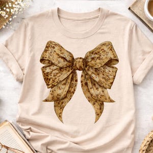 May include: A beige t-shirt with a large bow graphic. The bow is brown and tan with a spotted pattern. The t-shirt has a crew neck and long sleeves. The bow's design gives it a textured appearance.