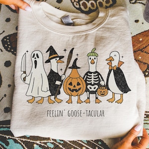 May include: Beige sweatshirt featuring a Halloween-themed graphic. The design includes five cartoon geese dressed in costumes: a ghost, a witch, a pumpkin, a skeleton, and a vampire. The text "FEELIN' GOOSE-TACULAR" is printed below the geese.