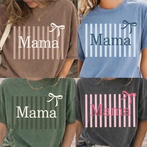 Könnte beinhalten: Vier kurzärmelige T-Shirts in Braun, Blau, Grün und Grau. Jedes Shirt zeigt das Wort "Mama" in Weiß, mit vertikalen Streifen und einem Schleifendesign. Die Shirts sind Freizeitkleidung.