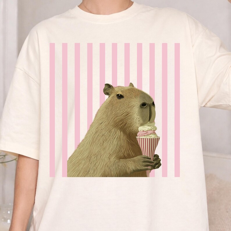 May include: Cream-colored t-shirt featuring a capybara enjoying an ice cream cone. The capybara is light brown with a detailed fur texture. The background has pink and white vertical stripes.