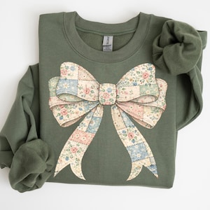 Könnte beinhalten: Ein olivgrünes Sweatshirt mit einem großen Schleifendesign. Die Schleife hat ein Patchworkmuster mit floralen und Pastellfarben, darunter Rosa, Blau und Creme. Das Sweatshirt hat einen Rundhalsausschnitt und lange Ärmel.