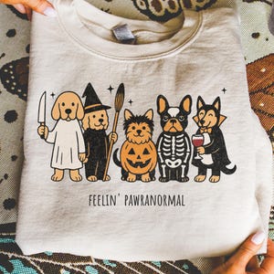 Halloween PNG for download - funny dogs in halloween costumes with quote feelin pawranormal. Halloween-themed graphic of five cartoon dogs dressed in costumes. The dogs are dressed as a ghost, witch, jack-o'-lantern, skeleton, and vampire. The text "FEELIN' PAWRANORMAL" is printed below the dogs.