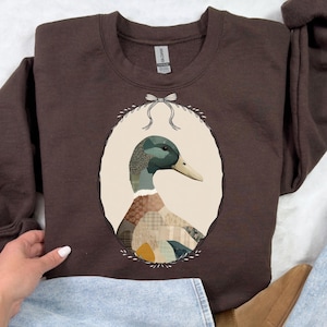 May include: A brown sweatshirt featuring an oval portrait of a duck. The duck has a green head, a tan beak, and a patchwork design on its chest. The oval is framed by a thin black line and a bow at the top.