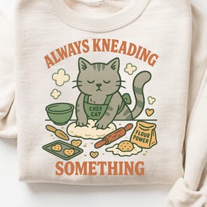 Puede incluir: Sudadera color crema con un gato gris con delantal verde que dice "Chef Cat". El gato amasa masa. El texto dice "Always Kneading Something" y "Flour Power". Diseño de panadería.