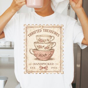 K&ouml;nnte beinhalten: Wei&szlig;es T-Shirt mit einem Vintage-Grafikdesign von drei gestapelten Teetassen mit floralen Mustern. Das Design enth&auml;lt den Text "THRIFTED TREASURES", "HANDPICKED" und "EST. 1956" innerhalb eines Briefmarkenrahmens.