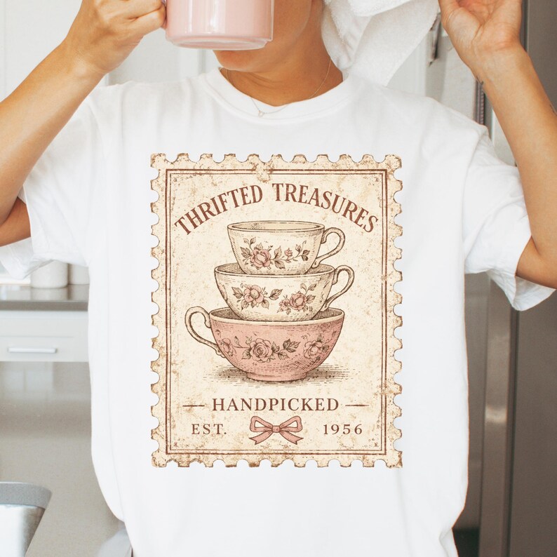 K&ouml;nnte beinhalten: Wei&szlig;es T-Shirt mit einem Vintage-Grafikdesign von drei gestapelten Teetassen mit floralen Mustern. Das Design enth&auml;lt den Text "THRIFTED TREASURES", "HANDPICKED" und "EST. 1956" innerhalb eines Briefmarkenrahmens.