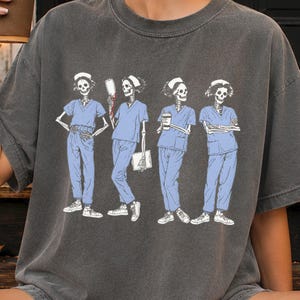 Può includere: Una t-shirt grigio antracite con una stampa grafica di quattro scheletri che indossano divise mediche azzurre e cuffiette da infermiere bianche. Gli scheletri sono raffigurati in varie pose, con uno che regge una sacca per flebo, uno una tazza di caffè e uno una cartellina, il tutto in una palette di colori bianco e blu.