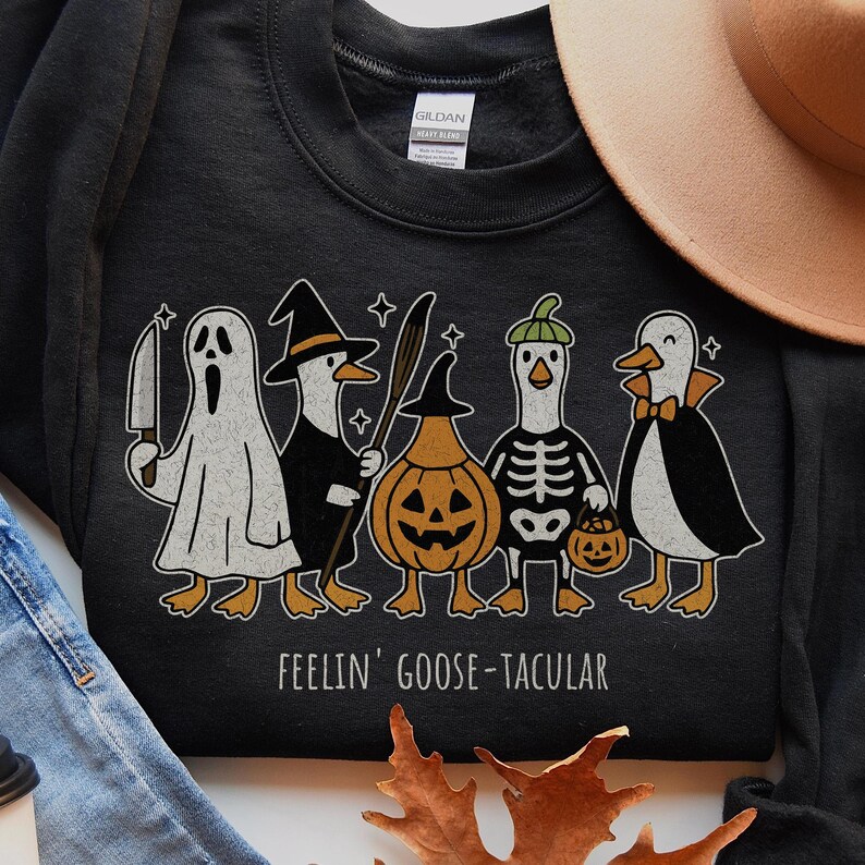 May include: Black sweatshirt with a Halloween-themed graphic. The design features five cartoon geese dressed in costumes: a ghost, a witch, a pumpkin, a skeleton, and a penguin. The text "FEELIN' GOOSE-TACULAR" is printed below the geese.