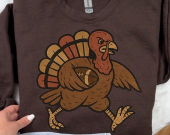 Könnte beinhalten: Braunes Sweatshirt mit einem Cartoon-Truthahn, der einen Football hält. Der Truthahn hat einen roten Kopf, braune Federn und einen bunten Schwanz. Das Design ist im Vintage-Stil, passend für Thanksgiving oder Football-Fans.