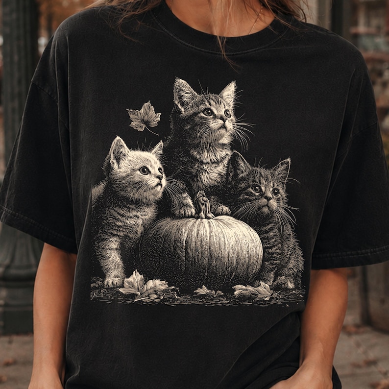 May include: A black short-sleeve t-shirt featuring a graphic print of three kittens sitting on and around a pumpkin. The kittens are depicted in a detailed, monochromatic style, with one kitten perched on top of the pumpkin and two others beside it.