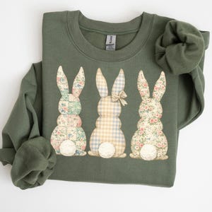 Könnte beinhalten: Olivgrünes Sweatshirt mit drei Hasen-Applikationen. Die Hasen haben Blumen- und Karomuster mit weißen, flauschigen Schwänzen. Ein Hase hat eine Schleife. Das Sweatshirt ist ein bequemer, lässiger Stil.
