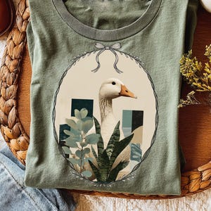 Puede incluir: Camiseta verde salvia con una ilustración detallada de un ganso blanco con pico naranja, rodeado de plantas estilizadas y formas geométricas dentro de un marco ovalado. El diseño incluye un lazo en la parte superior. La camiseta es de material suave.