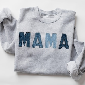 Peut inclure: Un sweat-shirt gris avec le mot "MAMA" sur le devant. Les lettres sont en tissu denim de différentes nuances de bleu. Le sweat-shirt a un col rond et des manches longues avec des poignets côtelés.
