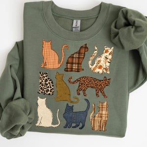 May include: An olive green crewneck sweatshirt featuring a graphic print of nine cats in various poses. The cats are filled with different patterns including orange plaid, brown plaid, floral, leopard print, solid tan, polka dot, and denim textures.