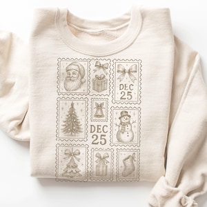 May include: Cream-colored sweatshirt featuring a collection of Christmas-themed postage stamp designs. The stamps depict Santa Claus, gifts, bows, a Christmas tree, a bell, a snowman, and a stocking. The date "DEC 25" is printed on two of the stamps.