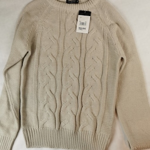 Peut inclure: Un pull en maille beige à torsades, à manches longues et à col rond. Le pull a une étiquette de prix indiquant "19,99€".