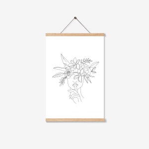 K&ouml;nnte beinhalten: Minimalistischer Kunstdruck mit einer Frau mit Blumenkranz, die einen Finger an den Lippen hat. Das Kunstwerk zeigt eine Geste der Stille. Das Bild ist auf wei&szlig;em Hintergrund und wird von einem Holzrahmen gehalten.