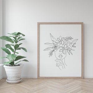 K&ouml;nnte beinhalten: Ein gerahmter Kunstdruck mit einer minimalistischen Strichzeichnung eines Gesichts mit Blumenkrone und einem Finger auf den Lippen. Das Schwarz-Wei&szlig;-Kunstwerk ist vor einem wei&szlig;en Hintergrund platziert und in hellem Holz gerahmt. Eine Topfpflanze befindet sich im Vordergrund.