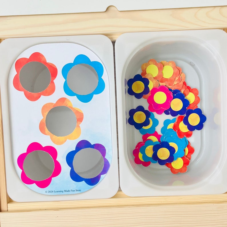 Op de afbeelding: Een kleurrijke bloemsorteeractiviteit voor kinderen. De activiteit omvat een ​​lade met vijf bloemvormige uitsnijdingen en een aparte container gevuld met kleurrijke bloemvormige stukken. De tekst "&copy; 2021 Learning Made Fun Store" is op de lade gedrukt.