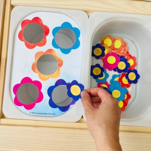 Op de afbeelding: Een kleurrijk bloemen-matchingspel met een witte achtergrond. Het spel bevat vijf bloemvormige uitsnijdingen met verschillende gekleurde centra en een container gevuld met bijpassende bloemvormen.