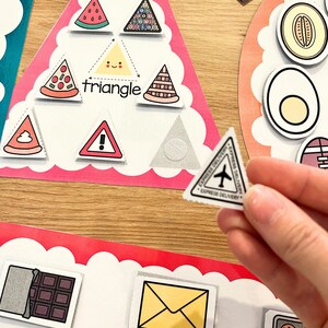 May include: A pink triangle-shaped learning card with various images, including a pizza slice, a watermelon, a triangle, a warning sign, a poop emoji, and a small airplane. The word "triangle" is written on the card.