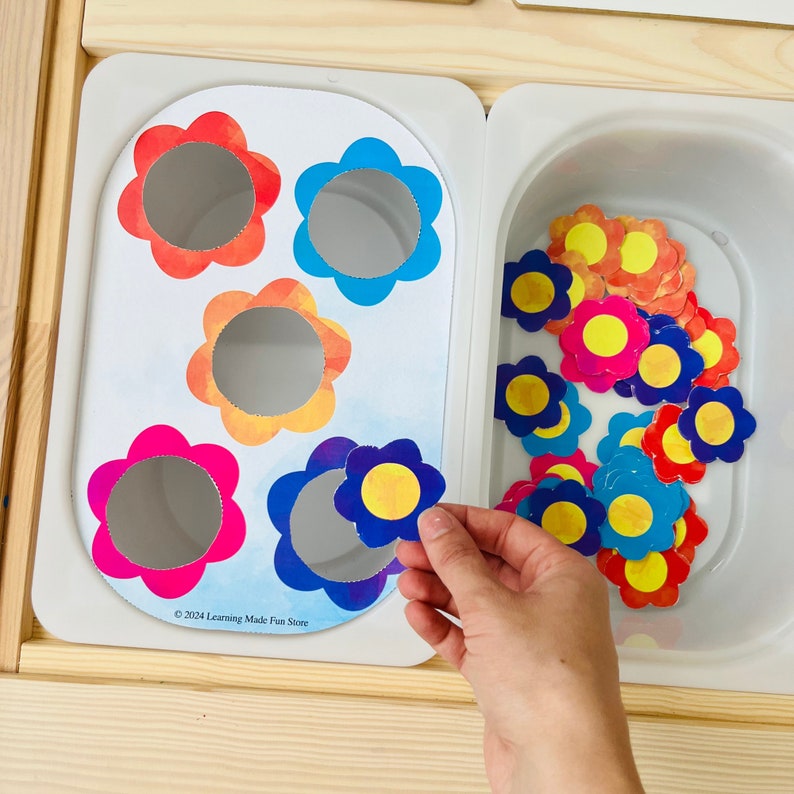 Op de afbeelding: Een wit bord met vijf kleurrijke bloemvormige uitsnijdingen. De uitsnijdingen hebben gaten in het midden. Een hand houdt een blauwe bloemuitsnijding vast met een geel midden. Een witte container met een verscheidenheid aan kleurrijke bloemuitsnijdingen bevindt zich aan de rechterkant van het bord. De tekst "&copy; 2024 Learning Made Fun Store" staat onderaan het bord.