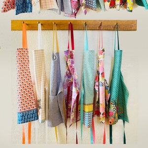 Peut inclure: Une collection de 13 torchons et tabliers color&eacute;s accroch&eacute;s &agrave; un support en bois. Les torchons et tabliers pr&eacute;sentent divers motifs et designs, notamment floraux, g&eacute;om&eacute;triques et &agrave; pois. Certains torchons portent un texte imprim&eacute;, comme "Wish Upon a Star".