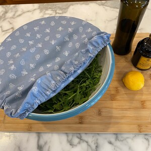May include: A light blue bowl cover with a white insect pattern, covering a bowl of rocket. The cover has a ruffled edge. Beside the bowl are a bottle of olive oil, a bottle of balsamic vinegar, and a lemon on a wooden chopping board.