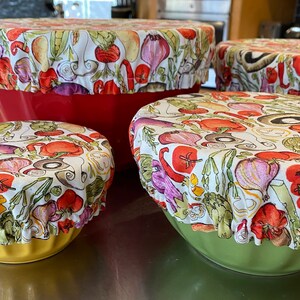 May include: A collection of four bowl covers, each featuring a fabric design with a white backdrop and a pattern of vegetables in red, purple, green, and yellow. The covers are placed on top of red, yellow, and green bowls.
