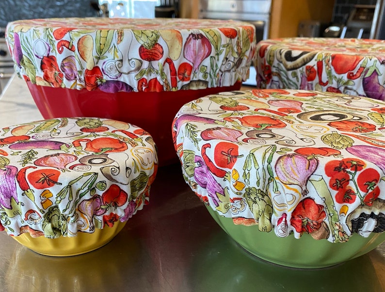 May include: A collection of four bowl covers, each featuring a fabric design with a white backdrop and a pattern of vegetables in red, purple, green, and yellow. The covers are placed on top of red, yellow, and green bowls.