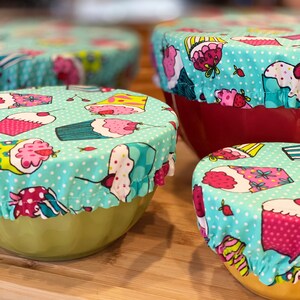 May include: Four bowls with cupcake-patterned fabric covers. The covers are light blue with white polka dots and feature various cupcake designs in pink, yellow, and green. The bowls are in shades of red and green.