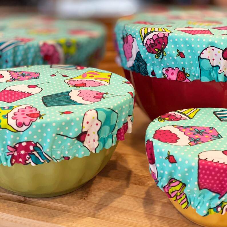 May include: Four bowls with cupcake-patterned fabric covers. The covers are light blue with white polka dots and feature various cupcake designs in pink, yellow, and green. The bowls are in shades of red and green.