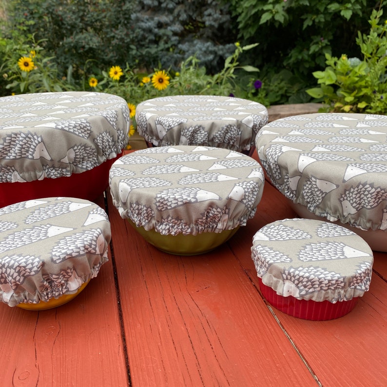 May include: Six fabric bowl covers with a repeating pattern of hedgehogs in grey and white. The covers are placed over bowls of different sizes and colours, including red, yellow, and green. The bowls are on a red wooden surface.