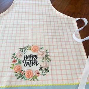 Könnte beinhalten: Eine rosa-weiße karierte Schürze mit blauem und gelbem Saum. Die Schürze hat eine gestickte Blumenkrone mit dem Text "Happy Easter" in Schwarz.