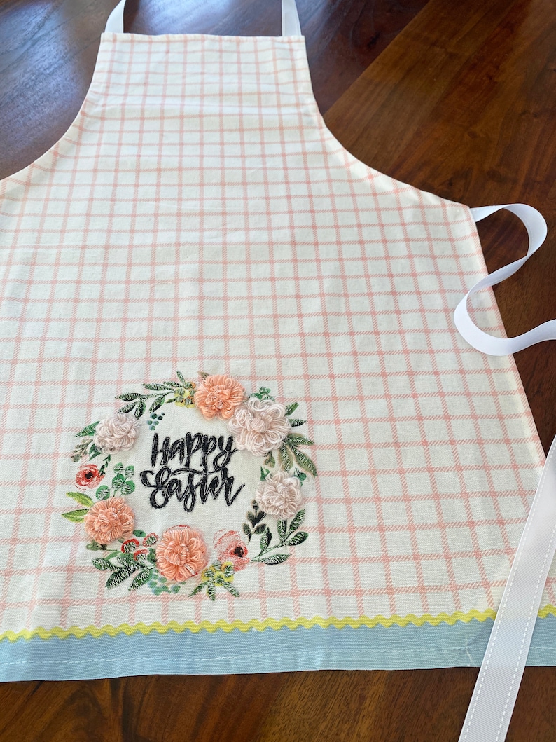 Peut inclure: Un tablier &agrave; carreaux rose et blanc avec une bordure bleue et jaune. Le tablier est orn&eacute; d'une couronne florale brod&eacute;e avec le texte "Happy Easter" en noir.