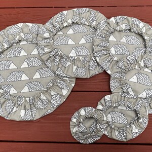 May include: A set of grey fabric bowl covers with elasticated edges. The covers feature a repeating pattern of white and grey hedgehogs. Various sizes are shown, suitable for different bowls. The covers are made of fabric.