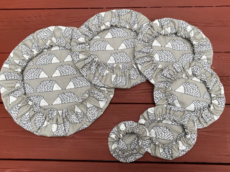 May include: A set of grey fabric bowl covers with elasticated edges. The covers feature a repeating pattern of white and grey hedgehogs. Various sizes are shown, suitable for different bowls. The covers are made of fabric.