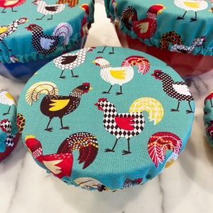 May include: A set of teal fabric bowl covers featuring a repeating rooster design in red, black, white, and yellow. The covers have ruffled edges and are placed on various coloured bowls, offering a charming and practical kitchen accessory.