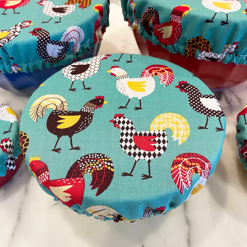 May include: A set of teal fabric bowl covers featuring a repeating rooster design in red, black, white, and yellow. The covers have ruffled edges and are placed on various coloured bowls, offering a charming and practical kitchen accessory.