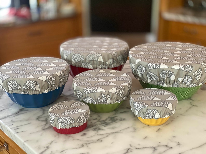 May include: A collection of six bowls with fabric covers. The covers are grey with a pattern of white and black hedgehogs. The bowls are in various colours, including blue, red, green, and yellow. The bowls are measured in centimetres.