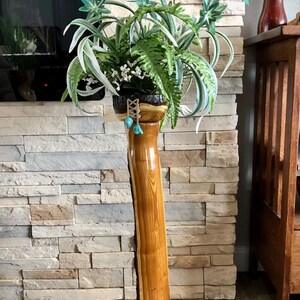 May include: A wooden plant stand with a natural wood finish. The stand is tall and slender, with a wide base and a narrow top. A green plant with blue flowers is in the pot on top of the stand.