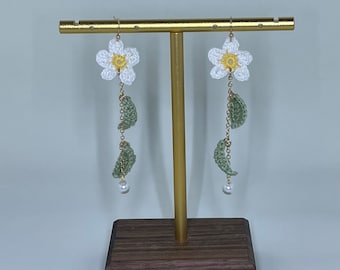May include: A pair of crocheted flower earrings with green leaves and pearl accents hanging from a gold stand.