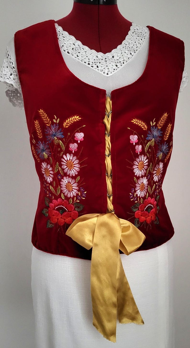 K&ouml;nnte beinhalten: Eine rote Samt-Weste mit gestickten Blumenmotiven in den Farben Rosa, Wei&szlig;, Blau und Gr&uuml;n. Die Weste hat ein goldenes Band, das an der Taille zu einer Schleife gebunden ist.