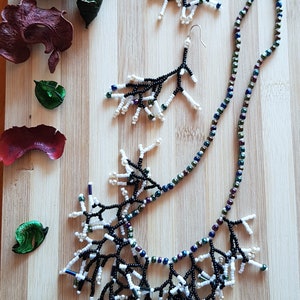 May include: A black and white beaded necklace with a branch-like design. The necklace is paired with matching earrings. The jewelry is displayed on a wooden surface.